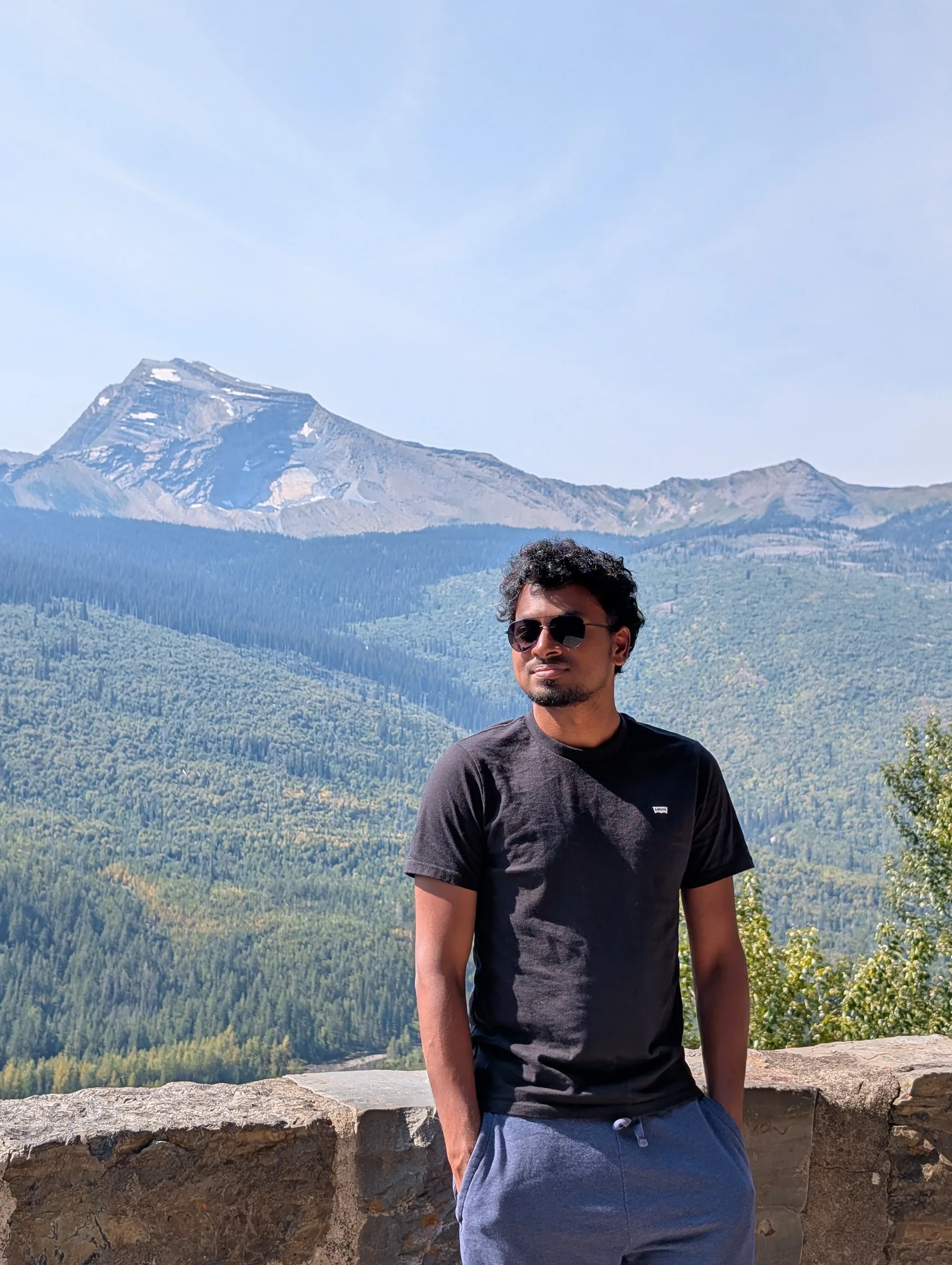Traveller at a stone overlook wall with forested valleys and a rocky peak behind, Glacier National Park