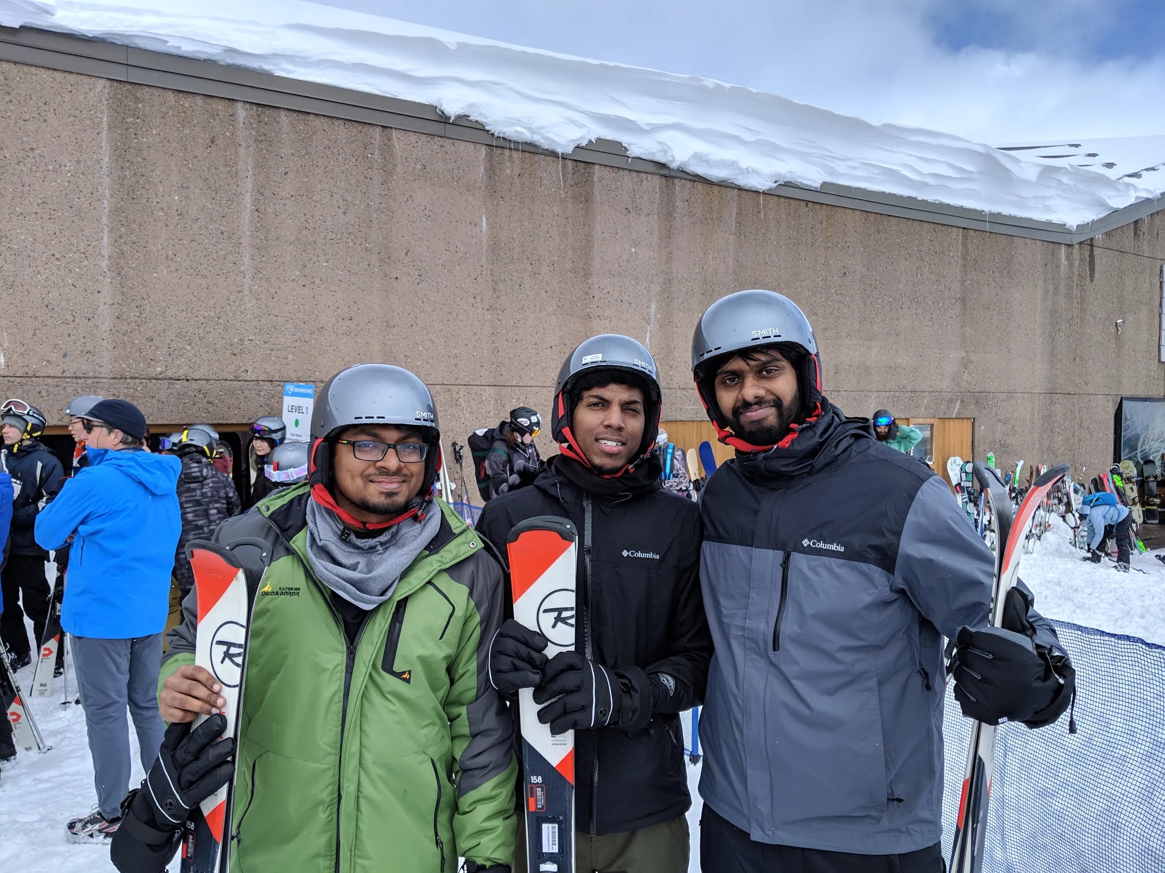 Three friends in helmets holding skis outside a snow-covered lodge on Mount Hood